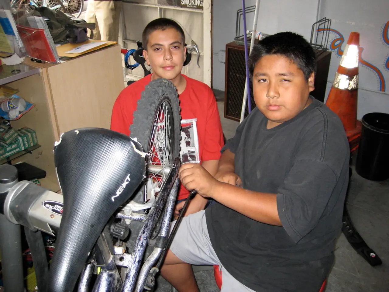 In the center of the picture there are two people repairing a bicycle. On the left there is desk,...